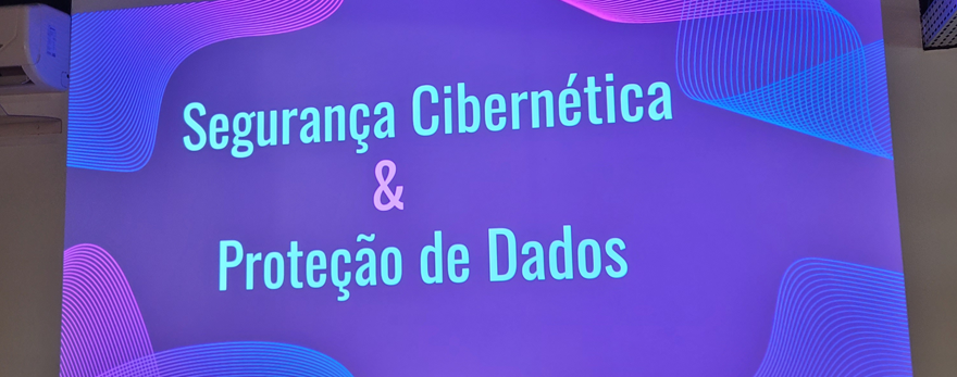 Fotografia ilustrativa: foto de uma arte gráfica projetada na parede. Ela tem fundo roxo, com detalhes em azul claro e rosa. No centro, em azul claro, está escrito "Segurança Cibernética & Proteção de Dados". Imagem do texto Como proteger os dados da sua empresa dos ataques cibernéticos?